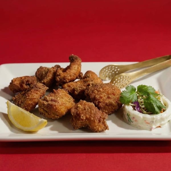 Crispy fried chicken pieces with a lemon wedge, coleslaw, and serving tongs on a white plate.
